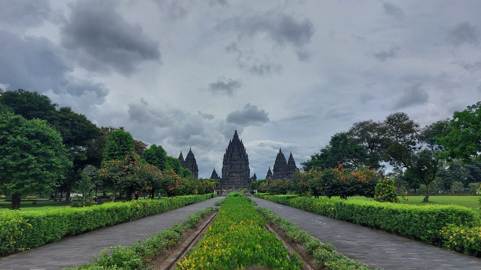 Kompleks świątynny Prambanan pod Yogyakartą — kamienne wieże na końcu parkowej alei pod zachmurzonym niebem